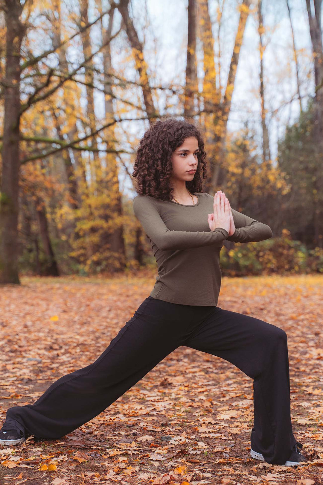 A woman practicing yoga in a forest with autumn leaves wearing an olive green long sleeve bamboo top and black wide leg yoga pants.