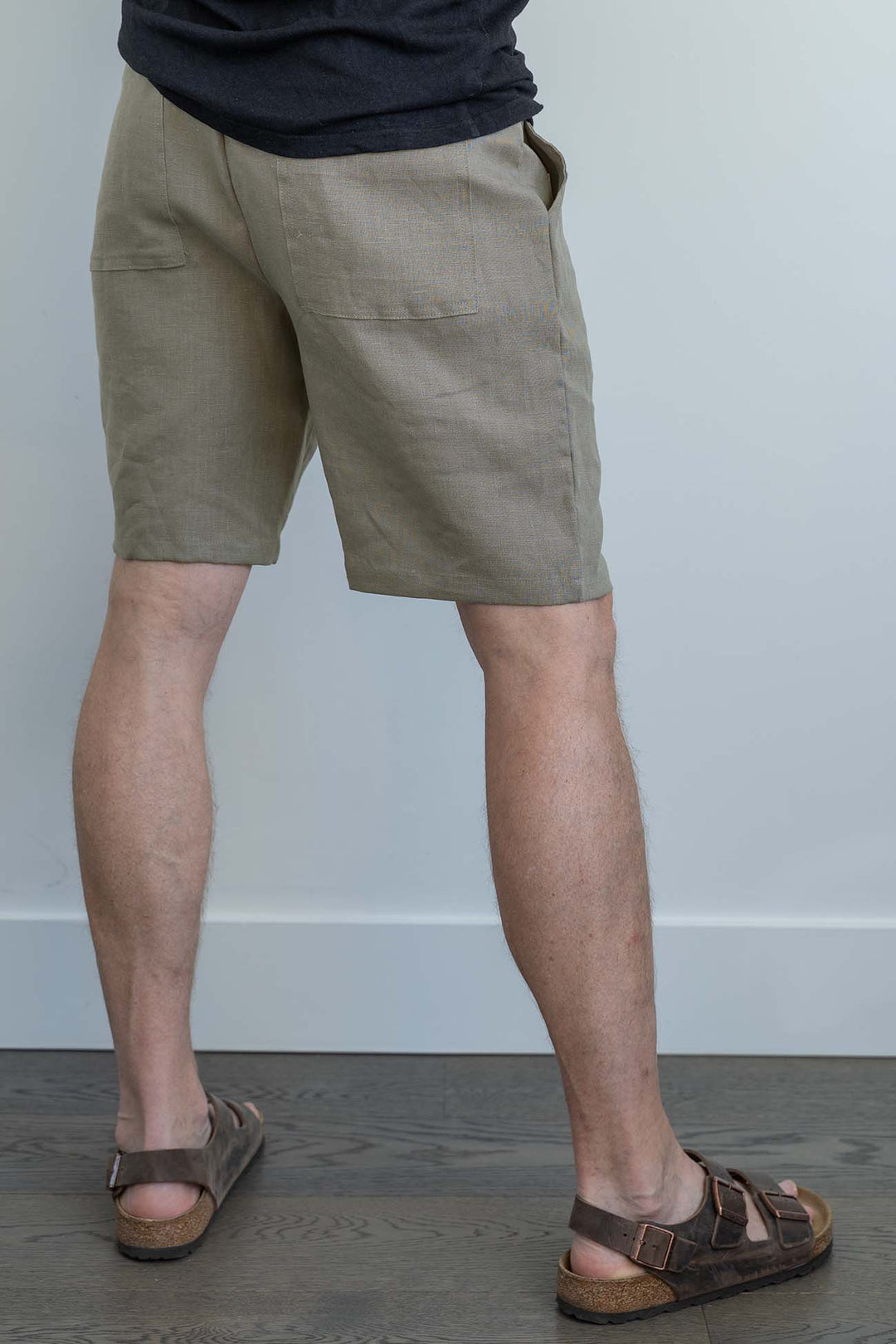A man standing facing a wall wearing taupe linen shorts with back pockets