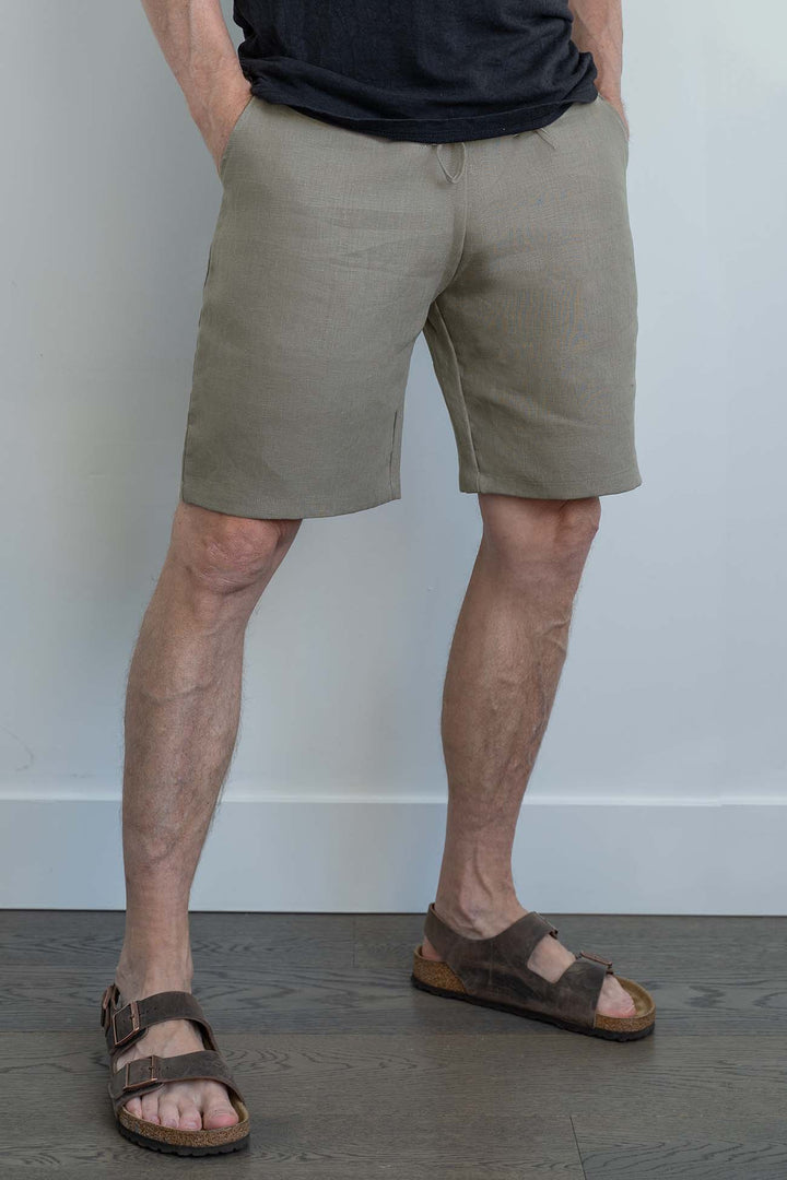 a man standing against a white wall wearing Taupe linen shorts and dark brown sandals