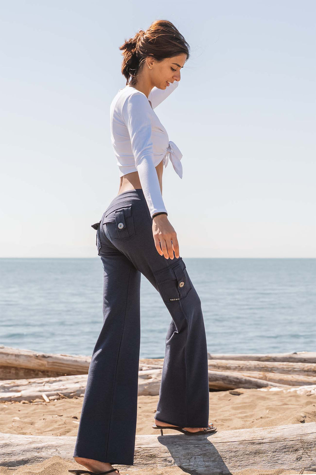 A woman stands on a sandy beach wearing dark navy wide-leg pocket cargo pants and a white tied long-sleeve crop top, with the ocean and sky in the background.