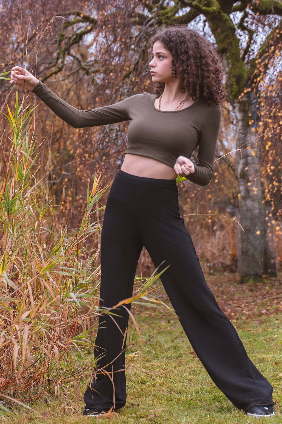Woman in an olive green bamboo long sleeve top and black high rise wide leg yoga pants standing outdoors with trees and grass in the background
