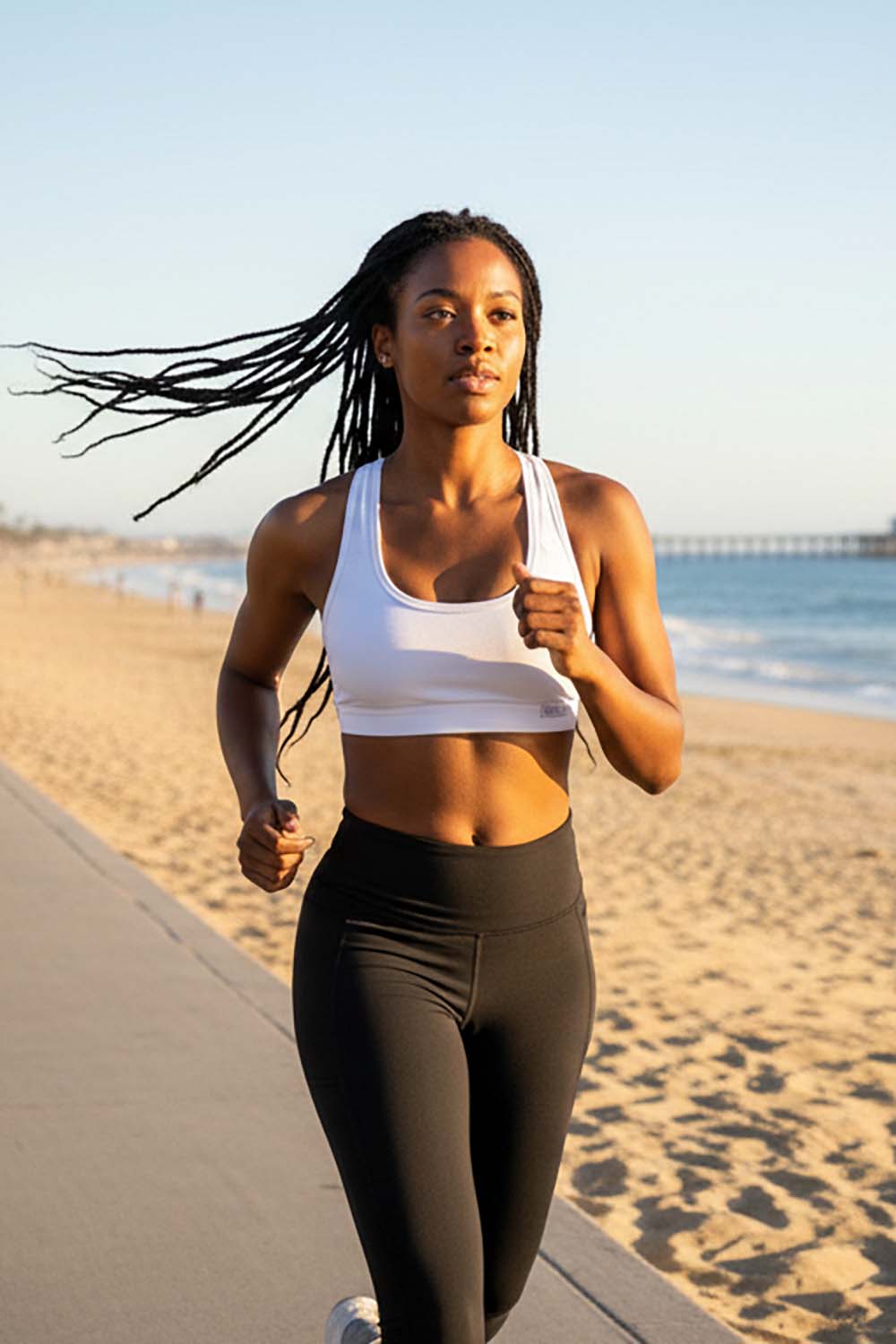 Woman wearing a white bamboo racerback sports bra and black leggings running in Manhattan beach  with ocean and sky in the background