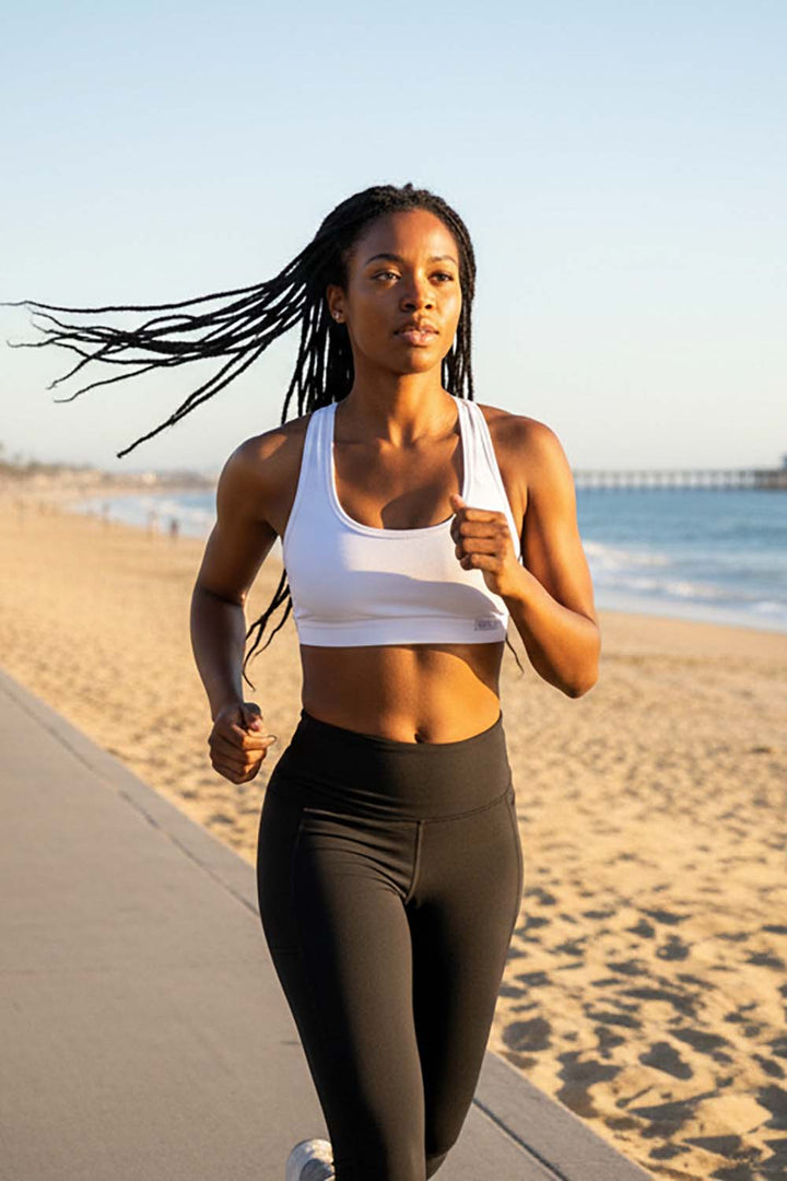 Woman wearing a white bamboo racerback sports bra and black leggings running in Manhattan beach  with ocean and sky in the background