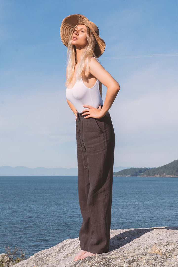 Woman in a white bamboo bodysuit and brown linen pants standing on a rock with a blue sky and water background