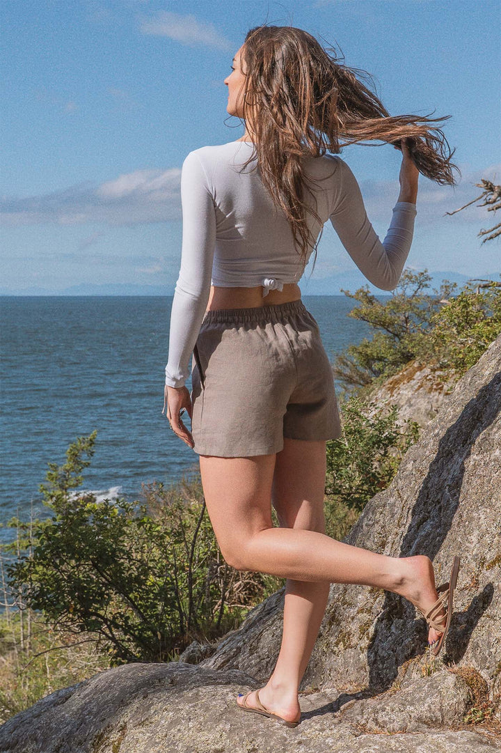 A woman wearing taupe colored linen shorts and a white long sleeve crop top standing on a rocking shoreline looking out over the ocean ion a windy day