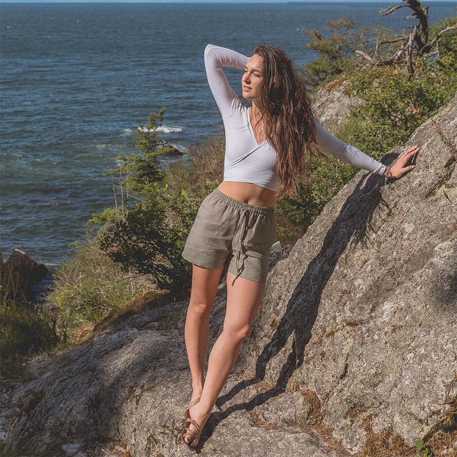 A woman standing on a rocky shoreline wearing taupe colored linen shorts and a white bamboo tie top with the open ocean in the background.